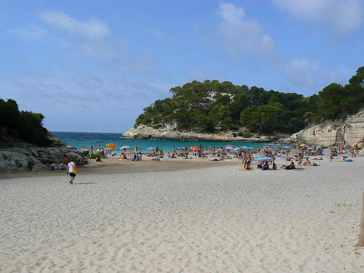 Cala Mitjana beach in Menorca packed with umbrellas in August, the cove a half-day boat loop reaches while the road car park is still filling
