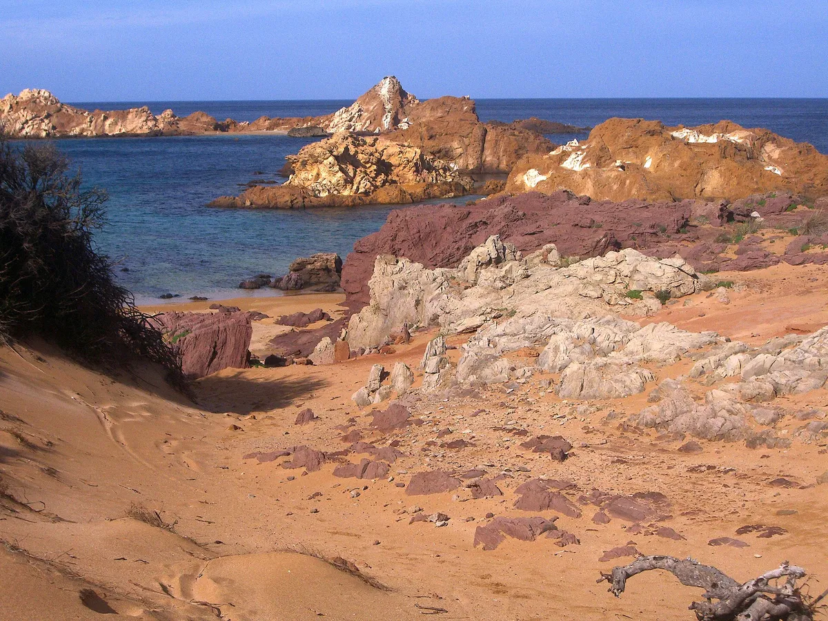 Red rocky coast and sheltered clear water at Cala Pregonda in northern Menorca seen from the dunes